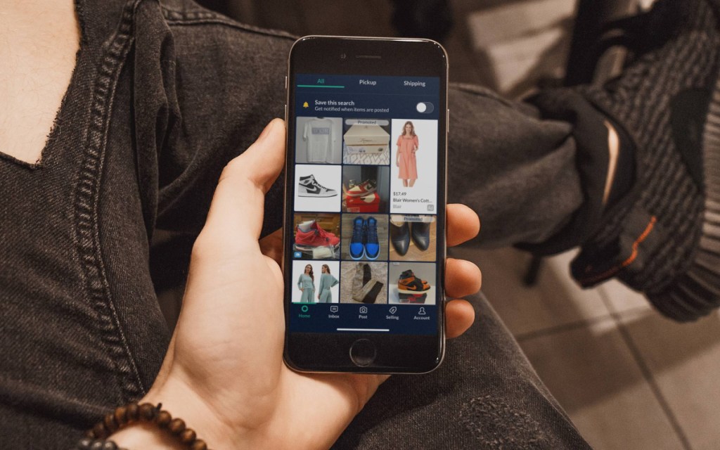 A hand holding a phone showing a marketplace selling app full of listings